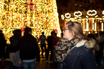 Blonde girl with glasses, looking at the Christmas lights of the city square. View from behind