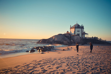 The chapel on the cliff on the oceanfront at sunset. People walk the beach, evening time. Capela do Senhor da Pedra in Miramar near Porto