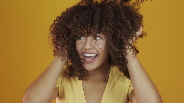 Happy laughing American African woman with her cyrly hair in yellow background. Laughing curly woman in sweater touching her hair and looking at the camera.