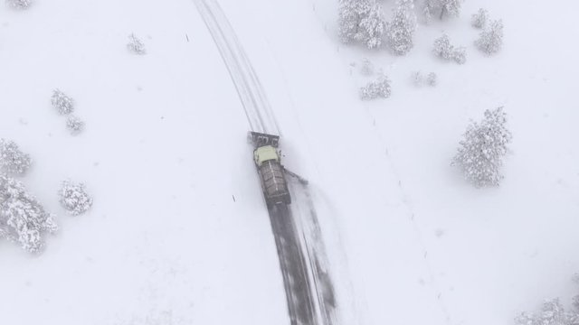 AERIAL: Flying above a snow plough truck clearing a snowy road during an intense snowstorm in rural America. Snow truck plows a rural highway in wintry Washington. Truck plowing and salting the road.