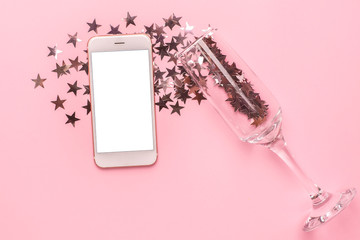 Mobile phone and champagne glass with silver stars confetti on pink color paper background minimal...