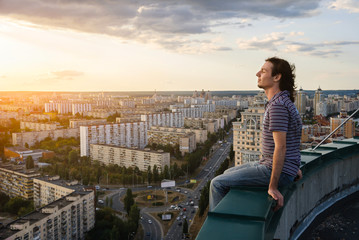 Young and brave man sitting on the edge of the roof and looking far away at the city, sunset...