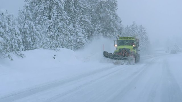 CLOSE UP: Truck plows the snowy country road during a horrible snowstorm raging in the state of Washington. Cars carefully drive behind a snow plough clearing the road during a devastating blizzard.