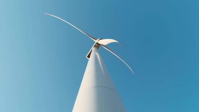 Slow motion of the blades of a large wind turbine in a field against a blue sky on a sunny summer day. View from the bottom up. Alternative energy sources. Windy park. Ecological energy. Industrial