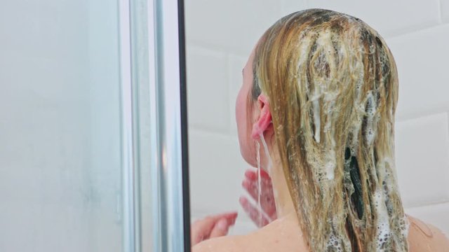 Back view of woman washing blond hair with shampoo in shower.