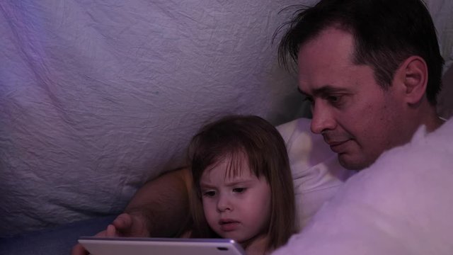 Father and daughter on Christmas evening, play and watch cartoons on tablet, in a children's room in tent with garlands. child and dad are playing in room. concept of happy childhood and family.