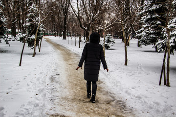 A girl in a black coat is walking along the road. Silhouette of a girl. Winter road. The girl in the hood in the winter. Black clothes. A winter day with trees and snow.