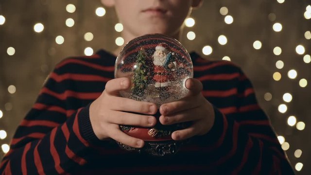 Christmas. Dreamy child holds a Christmas snow globe in his hands with Santa Claus, snow inside and makes Christmas present against the background of bright flickering bokeh lights. Boy Santa. Noel. 