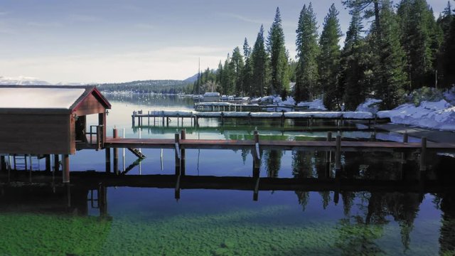 Aerial: flying over a pier and boat shed on a calm tranquil Lake Tahoe. Snow is piled up on the pier. Lake Tahoe, California, USA