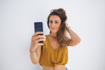 Smiling gorgeous lady in modern outfit capturing selfie photo by mobile on gray blank background
