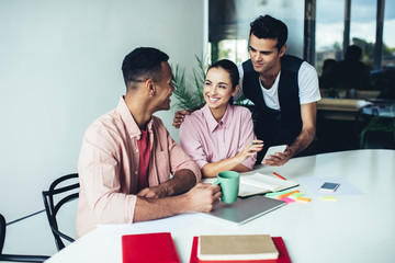 Happy male and female colleagues smiling during brainstorming meeting for creating ideas for startup project, prosperous multicultural group of friends talking near desktop using mobile phone