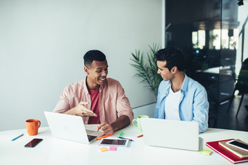 Cheerful male colleagues sitting at desktop with modern digital laptops and smiling during collaboration process, successful software developers discussing program code happy with brainstorming