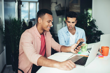 Multiracial office workers sitting at desktop with modern laptop computers and browsing websites for research information for startup, smiling men spending time for messaging via mobile application