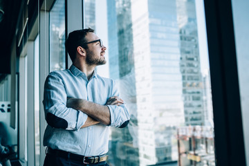 Contemplative male entrepreneur with crossed hands standing near office window view and feeling...