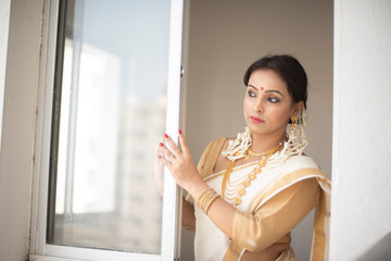 An young and attractive Indian woman in white traditional wear is smiling while standing in front of a glass window for the celebration of Onam/Pongal in white background. Indian lifestyle.