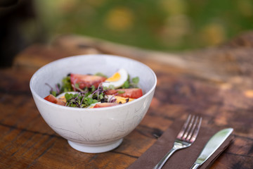 Healthy vegetable salad bowl mixed green leaves, eggs, black olives, tomato and bean sprouts on a wooden table with cutlery. Blurred outdoor background