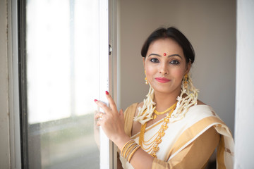 An young and attractive Indian woman in white traditional wear is smiling while standing in front of a glass window for the celebration of Onam/Pongal in white background. Indian lifestyle.