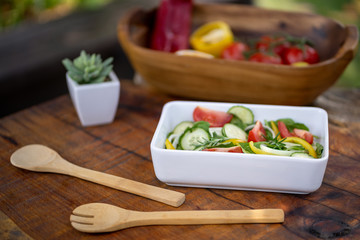 Salad with cucumber, baby spinach, onion and tomato served on a white plate on a wooden table. Behind there are wooden cutlery, a cactus and a bowl full of vegetables. Natural environment. Close up