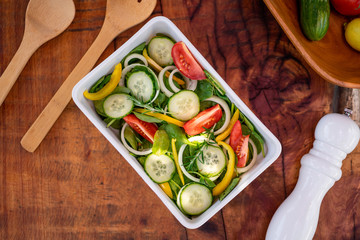 Fresh salad with cucumber, baby spinach, rosemary, onion and tomato served on a square white plate and wooden cutlery. Wooden background with a pepper shaker. Top view