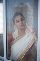 Blurred portrait of young and attractive Indian woman in white traditional sari and flowers is standing in front of closed window with water droplets/dewdrops on glass at Onam/Pongal.Indian lifestyle.