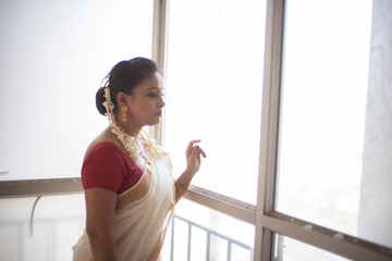 An young and attractive Indian woman in white traditional sari and red blouse and flowers standing in front of a glass window in a balcony  for the celebration of Onam/Pongal. Indian lifestyle.