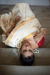 Top view of an young and attractive Indian  woman in white traditional wear is lying on a staircase in a festive day of Onam/Pongal in white background. Indian lifestyle.
