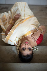 Top view of an young and attractive Indian  woman in white traditional wear is lying on a staircase in a festive day of Onam/Pongal in white background. Indian lifestyle.