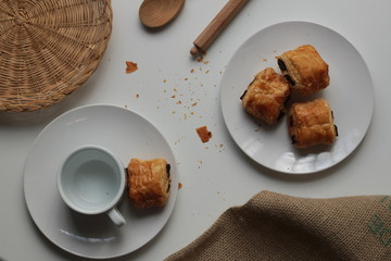 Flat lay of fresh, relaxed setting of black coffee and Pan Au Chocolate French inspired afternoon tea arrangement with clean white background with copy space and timber and brown accents and textures