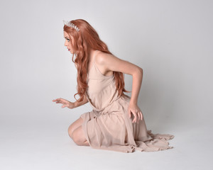 Fototapeta premium full length portrait of a pretty, fairy girl wearing a nude flowy dress and crystal crown. Seated pose against a grey studio background.