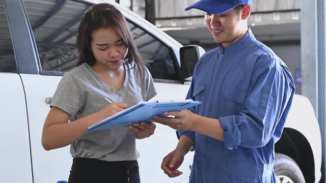 Asian car mechanic and female customer discussing to repairing and maintenance fixing automobile vehicle engine in auto repair shop garage background. Man and woman signing contract. Business concept.
