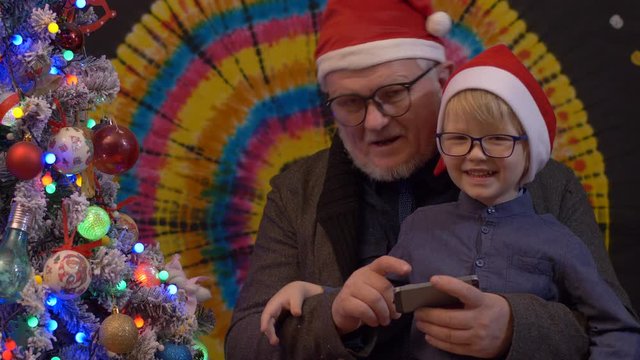 Father and son in glasses are sitting looking at images in smartphones near festively dressed Christmas tree. kid likes to play with his father in family holiday. New Year tree, colored background