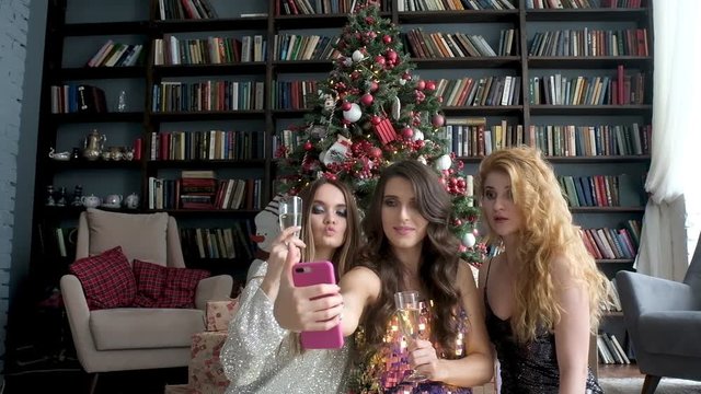 young women in evening dresses with glass of champagne take selfie near Christmas tree. Holiday with friends together. Christmas decorations and gifts. Balls and garlands on tree. Christmas mood