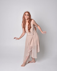 full length portrait of a pretty, fairy girl wearing a nude flowy dress and crystal crown. Standing and dancing pose against a grey studio background.