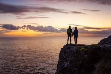 Adventurous couple holding hands and standing on a rocky cliff by the Pacific Ocean during colorful cloudy sunset. Taken in Lighthouse Park, West Vancouver, BC, Canada.