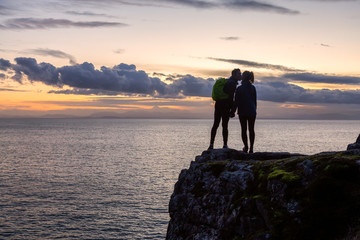 Adventurous couple holding hands and standing on a rocky cliff by the Pacific Ocean during colorful cloudy sunset. Taken in Lighthouse Park, West Vancouver, BC, Canada.