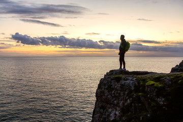 Adventurous Latino Male is standing on a rocky cliff by the Pacific Ocean during colorful cloudy...