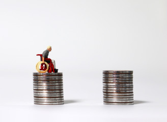 A miniature man in a wheelchair on a pile of coins next to a pile of coins.