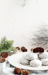 Chocolate cookies on a white plate on a background of fir cones and branches. Chocolate brownie cookies in white icing. Christmas gingerbread cookies. Holiday homemade baking.
