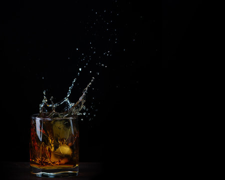 An ice cube falls into a glass with whiskey, leaving beautiful splashes on a black background