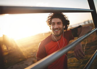 Portrait of a smiling young male athlete with earphones in his ears leaning on horizontal bars...