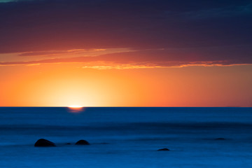 Vibrant colored summer sunset reflecting in ocean with endless horizon and deep blue ocean, silhouette of boulders laying in the foreground in shallow water at island of Gotland, Sweden