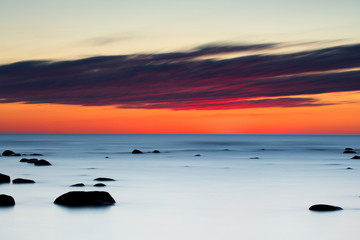 Fototapeta na wymiar Vibrant colored summer sunset reflecting in ocean with endless horizon and deep blue ocean, silhouette of boulders laying in the foreground in shallow water at island of Gotland, Sweden