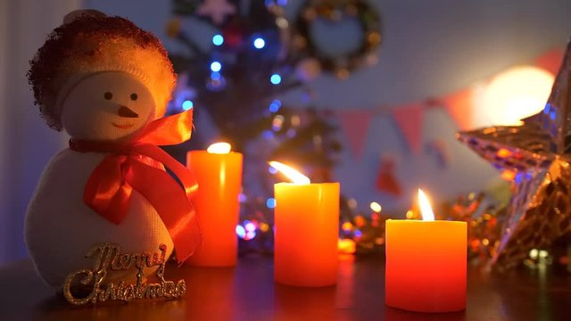 Closeup shot of a cute toy snowman with scented candles burning in a dark room - festive scene. Bokeh shot of a smiling snowman with a 'Merry Christmas' tag in a decorated room for Christmas celebr...