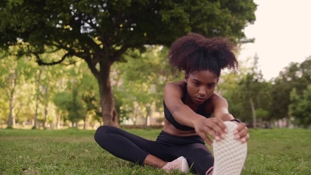 Healthy young african american woman in black sportswear stretching her legs before fitness exercises in the park