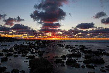 Fototapeta na wymiar Vibrant colored cloudy winter sunset reflecting in ocean with endless horizon and dark ocean, silhouette of boulders laying in the foreground in shallow water at island of Gotland, Sweden