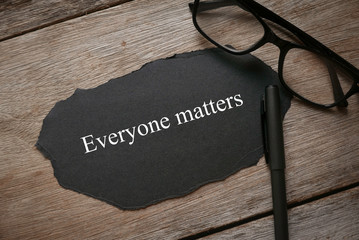 Glasses,pen and piece of black paper written with Everyone Matters on wooden background.