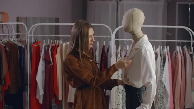 A beautiful girl in a jacket looks at clothes on a mannequin, touches and considers the style and type of fabric of clothes in the showroom