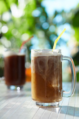 Subject shot of classic design beer mugs with dark refreshing drinks. The glass mugs with thick handles and coloured straws are situated on the white wood table in the summer outdoor interior. 