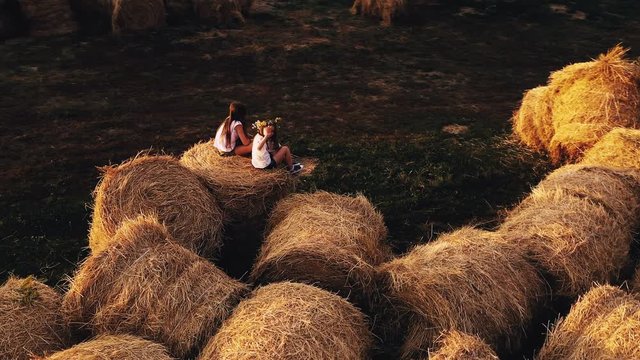 Two pretty girls sitting on massive haystacks in clear summer field, view from drone