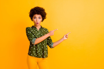 Photo of funny dark skin curly lady open mouth indicate fingers empty space show unbelievable low prices wear trendy green dotted shirt pants isolated yellow color background